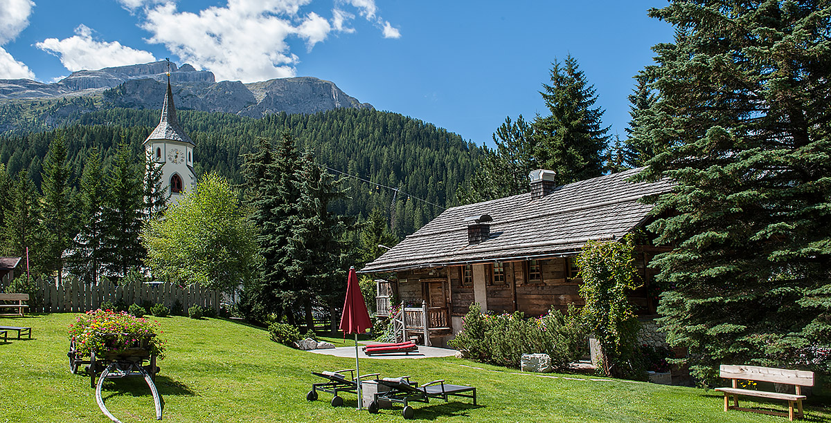 A wooden house in the woods and in the background, the bell tower of Corvara