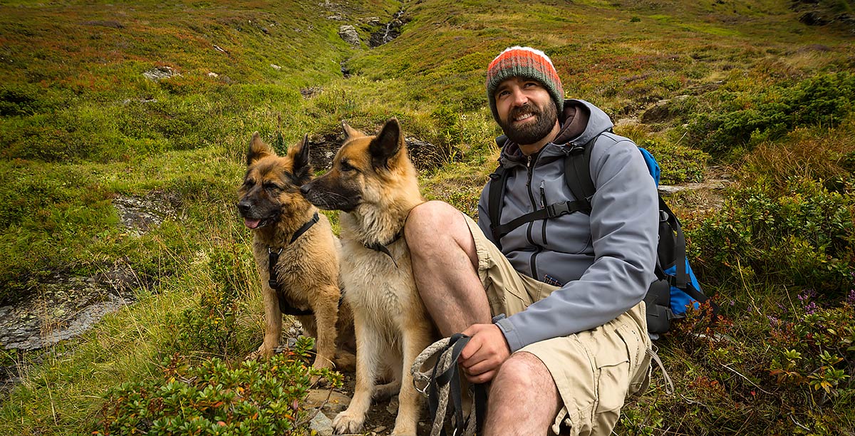 Man with backpack is having a break by sitting on a green mountain meadow with his two dogs.