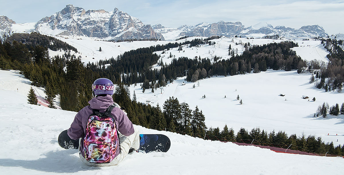 Mädchen mit Rucksack, Helm und Snowboard sitzt am Boden und beobachten die schneebedeckten Berge