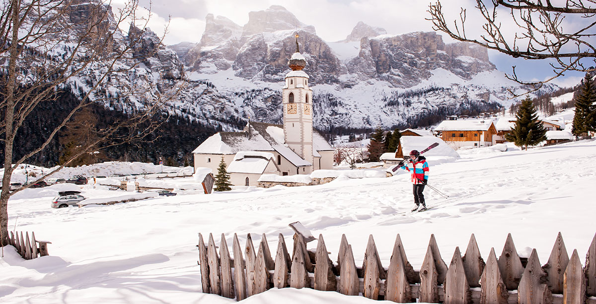 Ein Skifahrer im Vordergrund, der blaue Himmel, die Kirche und das Dorf von Colfosco im Hintergrund