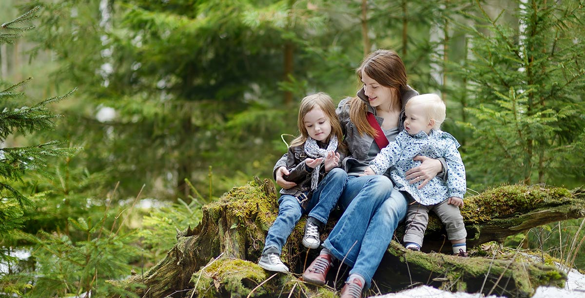 Mutter mit zwei Kindern auf dem Arm sitzt auf einem mit Moos bedeckten Baumstamm