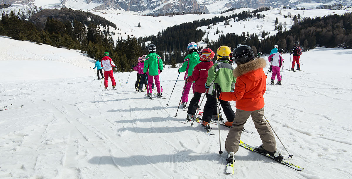 Children with skis in single file are preparing for a lesson with the teacher
